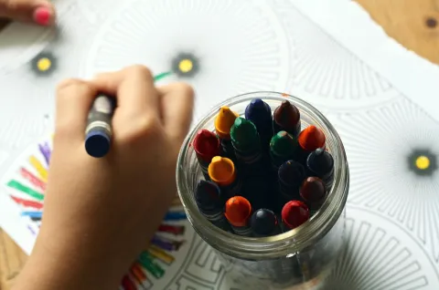 Coloring event.  A hand holding a crayon, coloring an image, next to a jar of crayons.