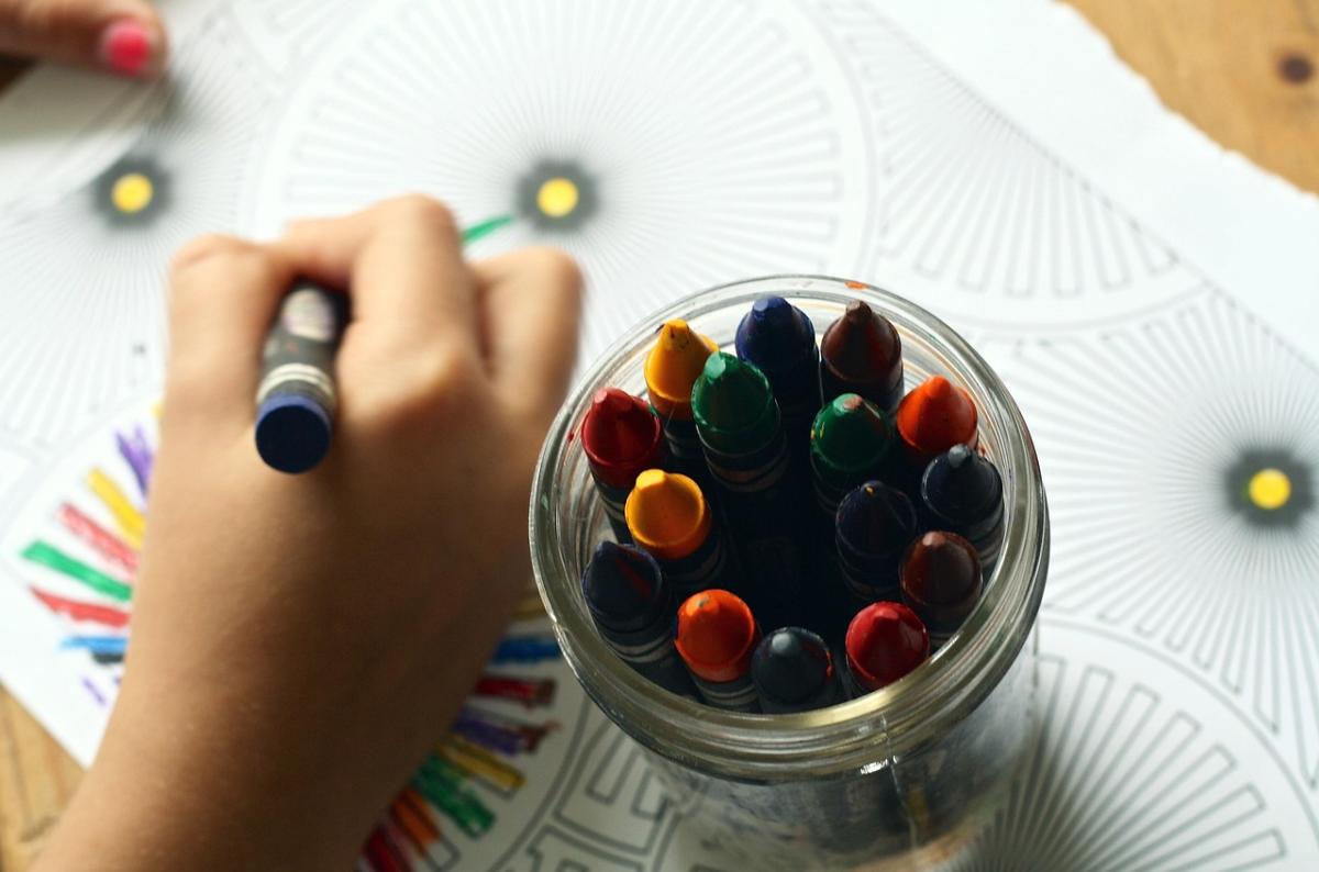 Coloring event.  A hand holding a crayon, coloring an image, next to a jar of crayons.
